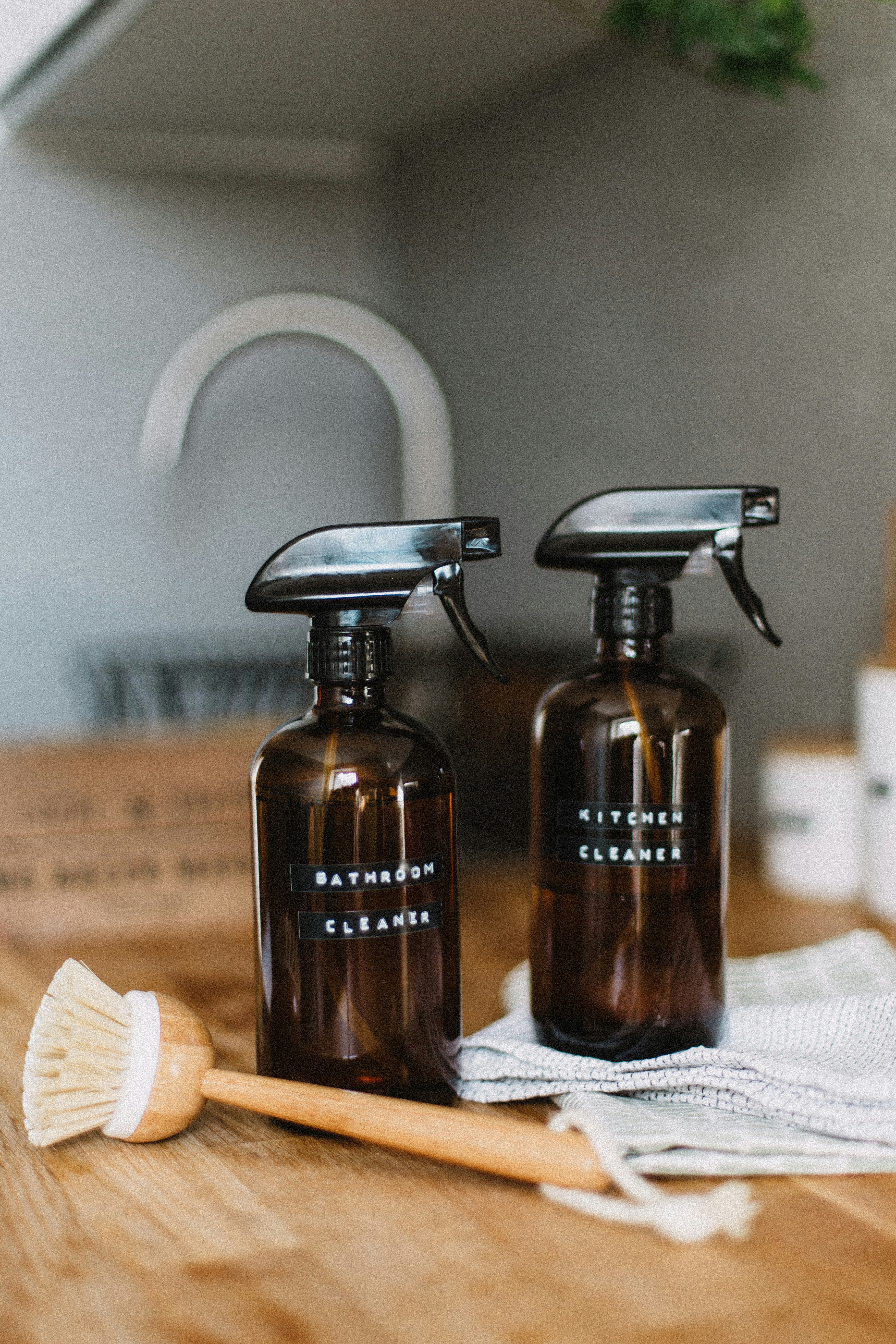 Two amber glass spray bottles labeled 'Bathroom Cleaner' and 'Kitchen Cleaner' on a wooden countertop, accompanied by a natural bristle scrub brush and a grey dish towel.